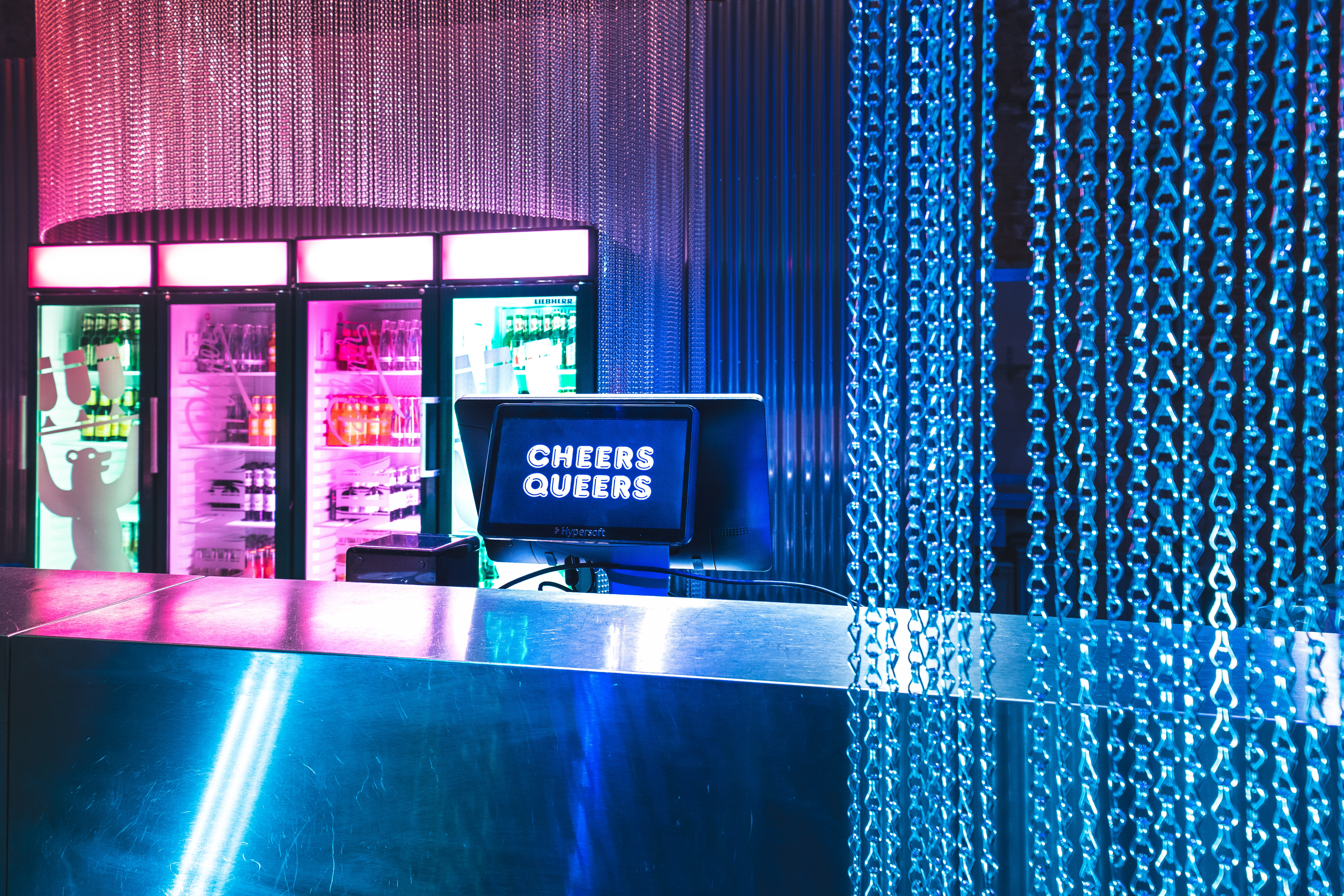 A bar in Schwuz with pink and blue lighting. The bar screen reads ‘Cheers Queers’.